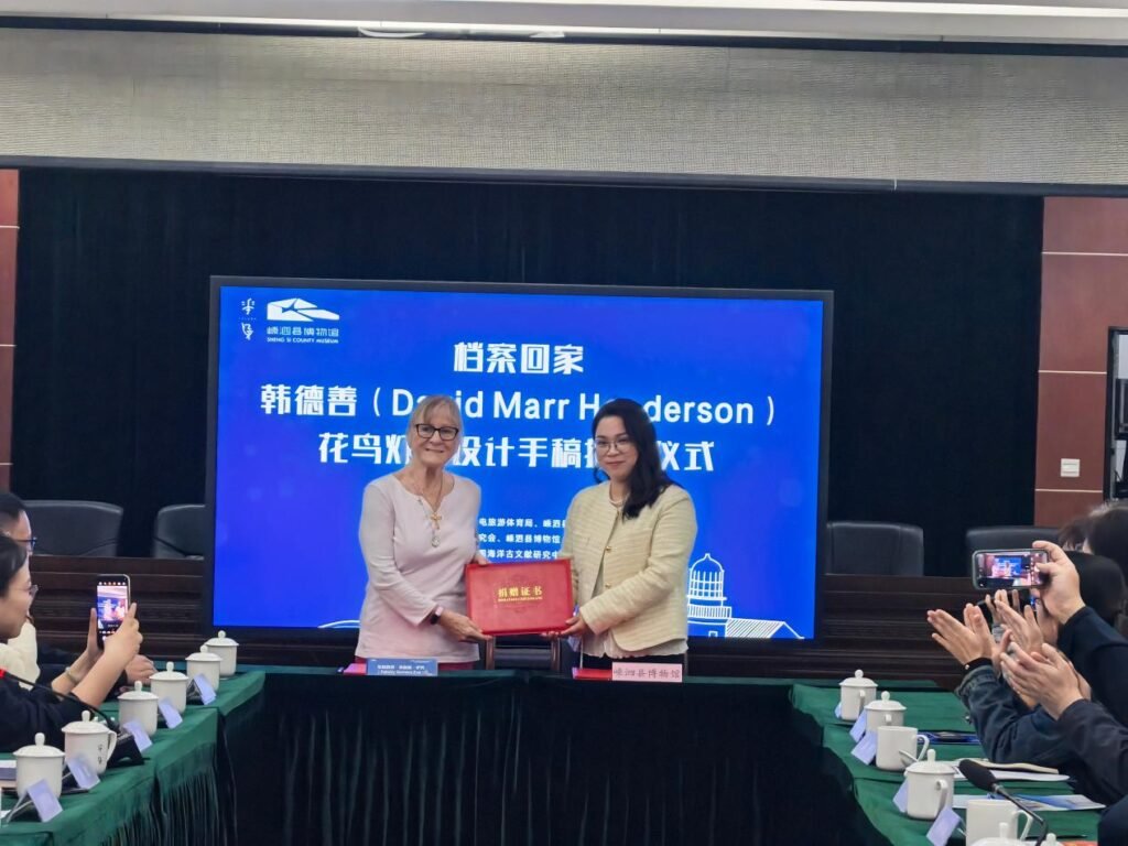 000234234234342342343423423443 - عروبة الاخباري | Oroba News Two women exchange a red folder at a formal signing ceremony in a conference room with a blue screen backdrop behind them.