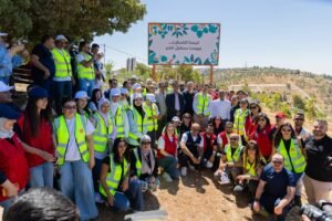 Diverse group of volunteers in high-visibility vests posing for a group photo outdoors beside a sign in Arabic.