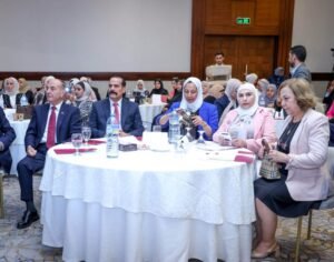A diverse group of professionals seated around round tables at a formal event in a conference hall, some taking notes and others using phones or cameras.