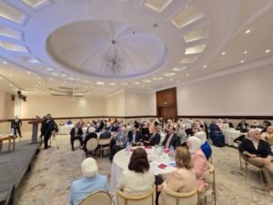 Banquet hall with round tables of formally dressed attendees listening to a speaker at a podium on a stage, chandeliers overhead and cameras recording.