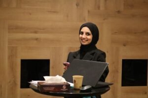 Smiling woman in a black hijab and blazer stands at a round table with a laptop, tissues, and a coffee cup in a wood-paneled room.