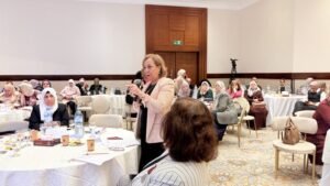 Woman standing at a round table speaking into a microphone during a conference with attendees in hijabs seated around tables.