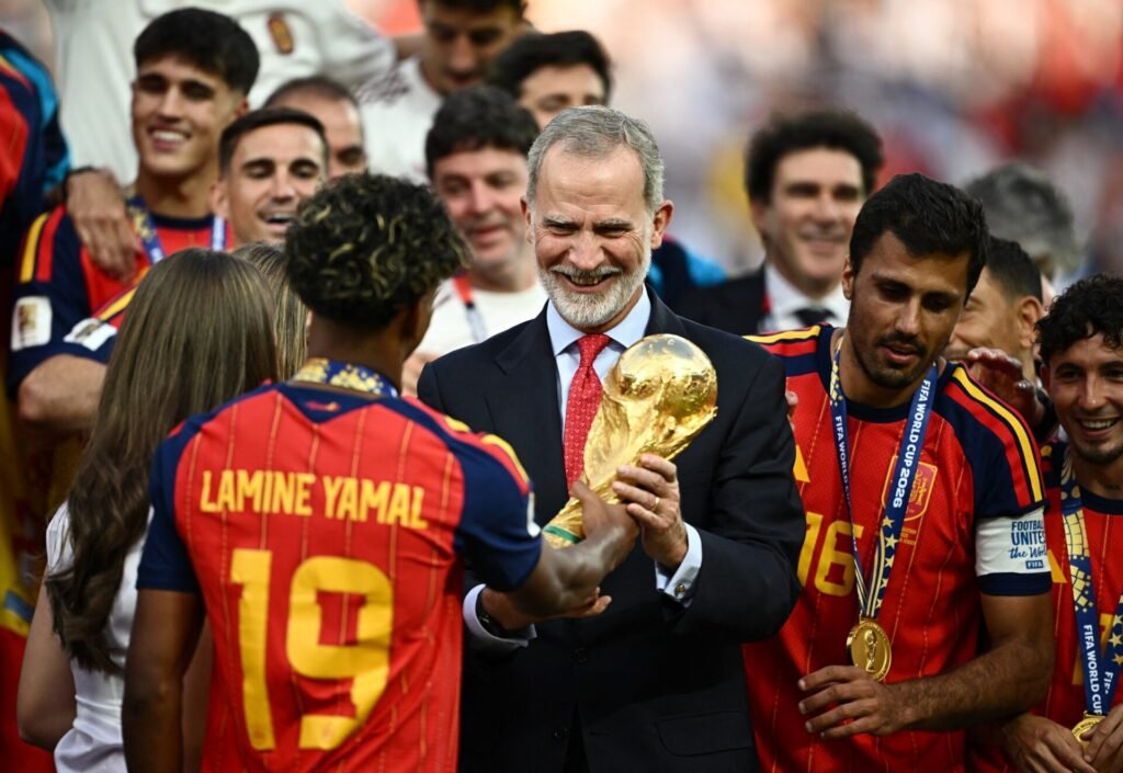 FIFA World Cup 2026 - Final - Spain v Argentina - عروبة الاخباري | Oroba News A man in a suit holds the FIFA World Cup trophy while teammates in red‑and‑yellow Spain jerseys celebrate around him on the field.