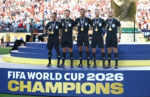 FIFA World Cup 2026 - Final - Spain v Argentina - عروبة الاخباري | Oroba News Five FIFA World Cup 2026 referees stand on a tiered podium wearing black uniforms and medals as a stadium crowd watches.