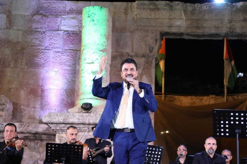 2027-24 at 50700 Male singer in a blue suit and yellow tie performing on an outdoor stage at night, with an orchestra behind him and illuminated stone walls behind.