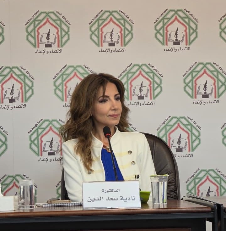 356422_0_1784174980 - عروبة الاخباري | Oroba News Dr. Nadia Saeed al-Din sits at a conference table with a microphone in front of her, backdrop repeating logo and Arabic text behind her, nameplate visible in front of her.