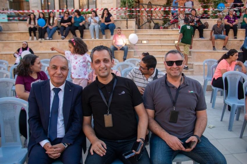 4005454 Three men sit in the front row at an outdoor event, wearing lanyards with badges, smiling for the photo in front of a crowd of seated attendees and kids in the background.