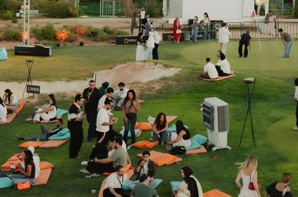 Group of people relaxing on colorful cushions and mats on a grassy outdoor lawn, chatting and socializing at an event.