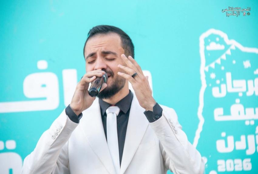 4009905454 Male singer in a white suit and black shirt passionately singing into a microphone on a bright turquoise stage backdrop with Arabic text.