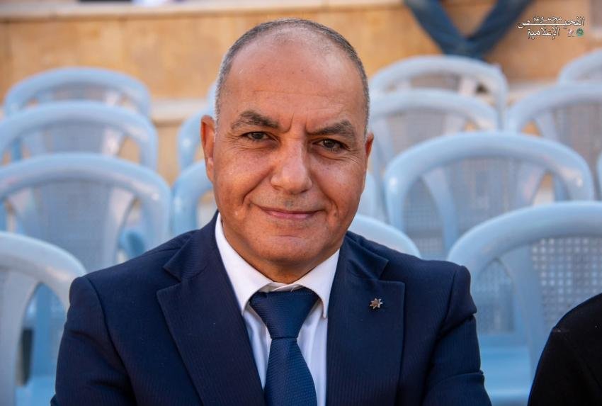 4009999905454 Smiling man in a dark navy suit and blue tie, seated outdoors with light blue plastic chairs in the background.