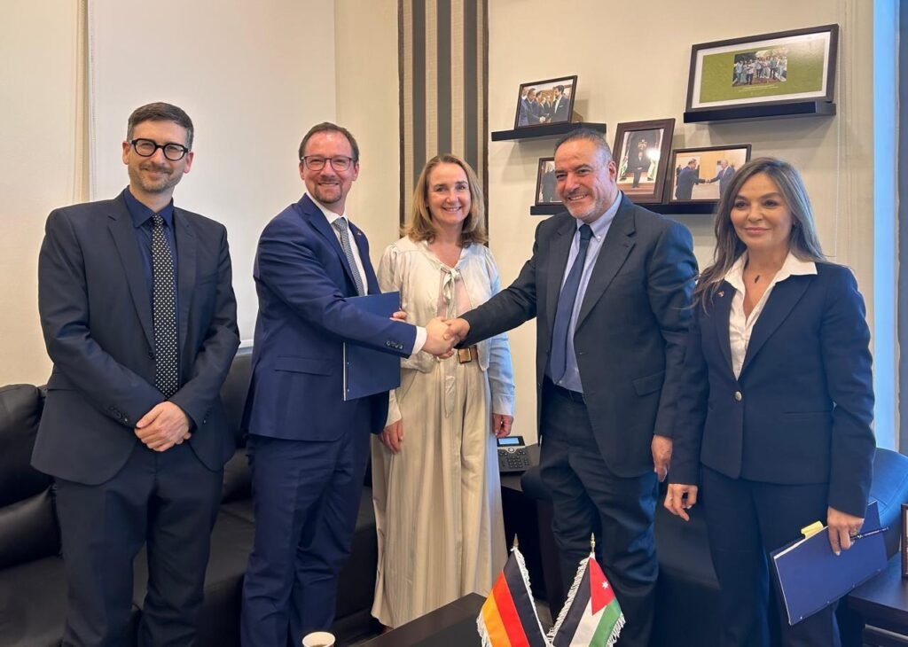 Five professionals in suits stand in an office while two men in the center shake hands, with two national flags on the table (Germany and Jordan) and framed photos on the wall behind them.