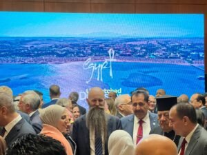 Crowd of people in formal attire at an indoor event, with a large blue screen showing a coastal aerial image in the background.
