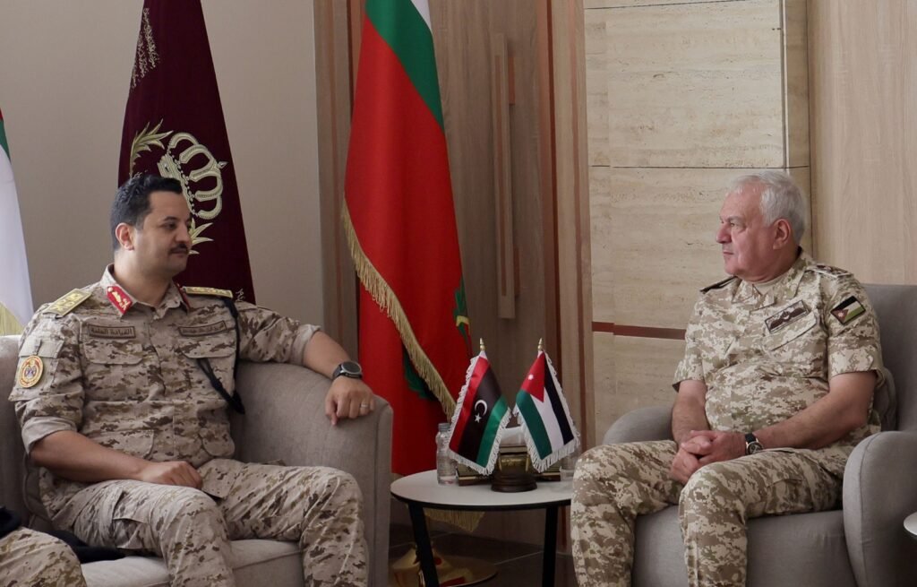 Two military officers in desert camouflage sit on sofas and talk in a formal meeting, with flags on a small table between them.
