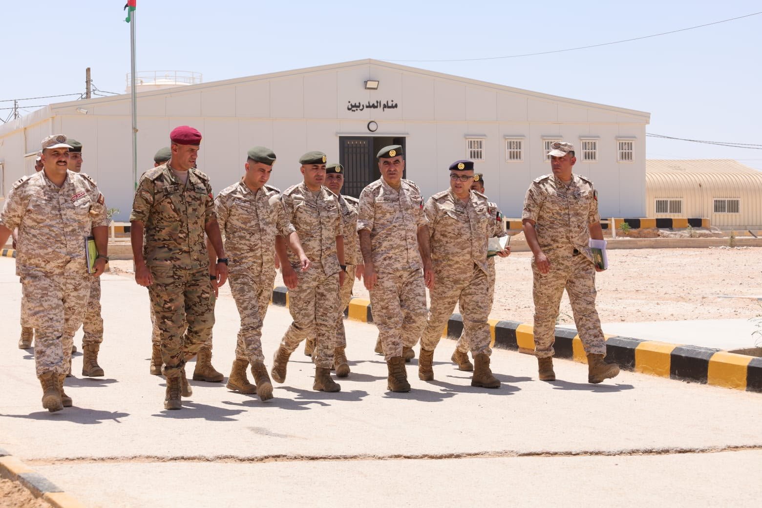 639202359137446571 A line of soldiers in desert camouflage walking together along a paved road outside a military building with Arabic signage on the facade.
