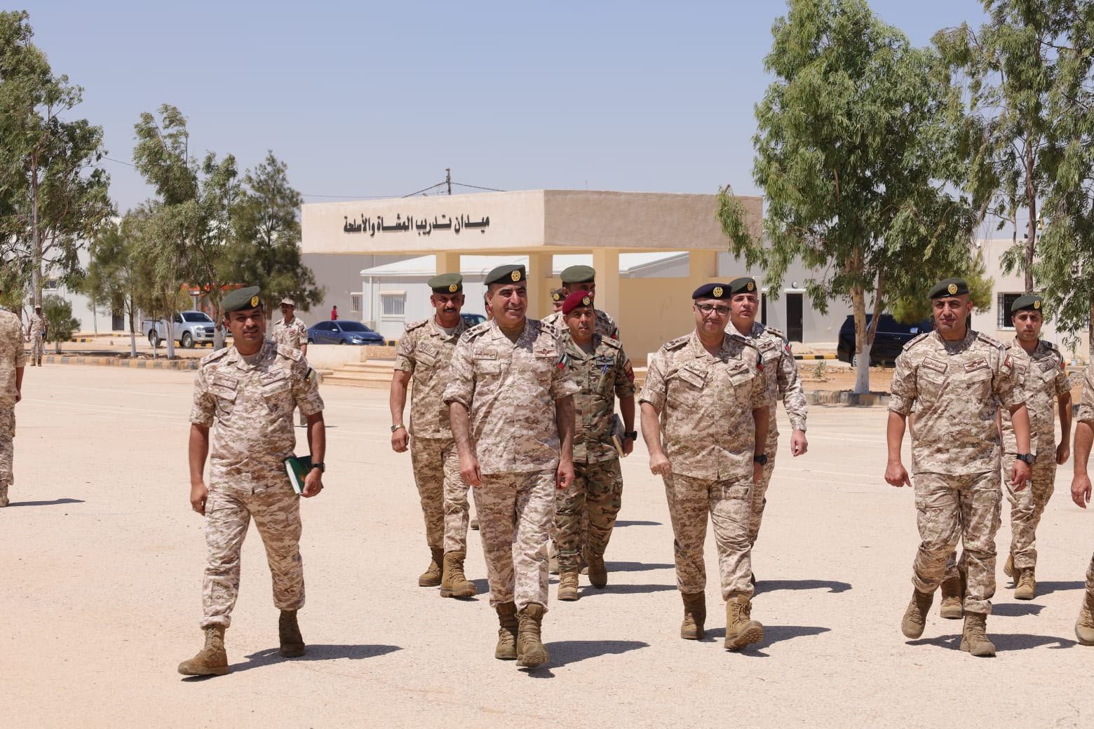 639202359138071610 Group of soldiers in desert camouflage walking across a sandy parade ground near a beige building with Arabic signage in the background.