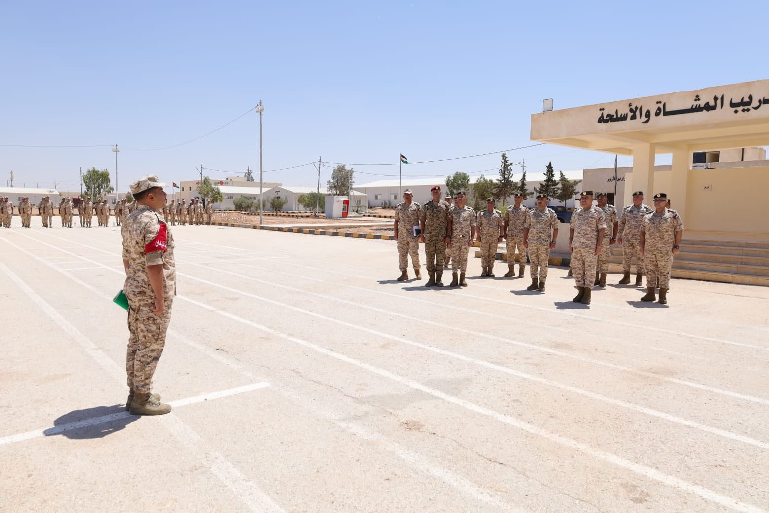639202359138852892 Commander facing a line of soldiers in desert camouflage on a sunny military base courtyard; building with Arabic signage in the background.