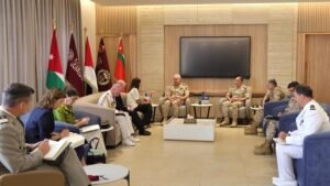 Formal meeting in a lounge with military officers and officials seated in a semicircle, flags in the background.
