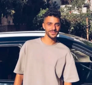 6a491c5465e3a - عروبة الاخباري | Oroba News Young man with short dark hair and a trimmed beard, in a beige T‑shirt, standing beside a black car outdoors.