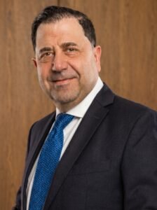 Professional headshot of a man in a dark suit and blue patterned tie, against a wood-paneled background, looking at the camera with a subtle smile.