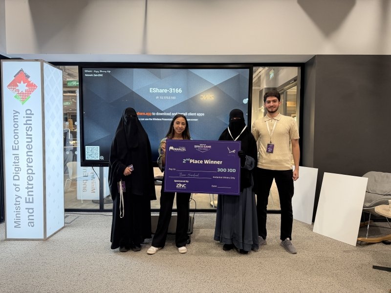 Group of four at a tech event: two women in abayas, a woman holding a large 2nd place prize check (300 JD); man on the right; Ministry banner on the left.