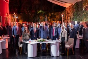 A group of formally dressed attendees stand behind banquet tables at an outdoor evening reception.