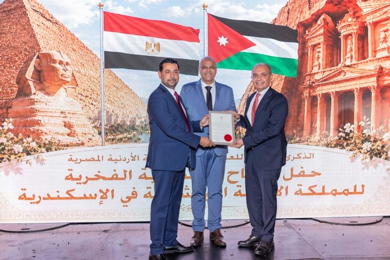 Three men in suits stand on a stage, presenting and receiving a framed certificate during an award ceremony with the Egyptian and Jordanian flags in the background.