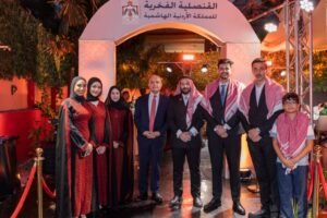 Group of nine adults and a child posing for a formal photo under an arched entryway with Arabic signage, nighttime event with suits and traditional scarves.
