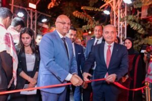 Group of formally dressed officials cutting a red ribbon at a ceremony, with onlookers and photographers nearby under bright event lighting