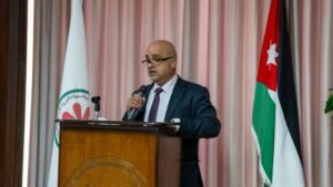 Man in a dark suit and purple tie speaks into a handheld microphone at a wooden podium, with a Jordanian flag to his right and a white banner with a red flower emblem to his left.