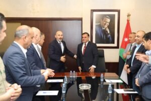 Officials in suits shake hands across a glass conference table during a formal meeting, Jordan flag and a framed portrait visible in the background.