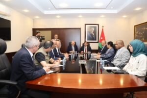 Group of professionals in a formal meeting around a wooden conference table; Jordan flag and a portrait are on the wall behind them.