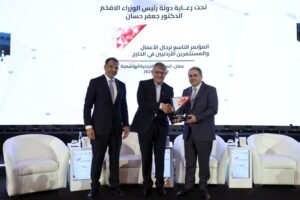 6a5dc1ac1accc - عروبة الاخباري | Oroba News ثلاثة رجال في بدلات رسمية على منصة حفل تكريم يتصافحون، بينما يُسلَّم أحدهم جائزة شفافة كرمز للبراءة والنجاح.
