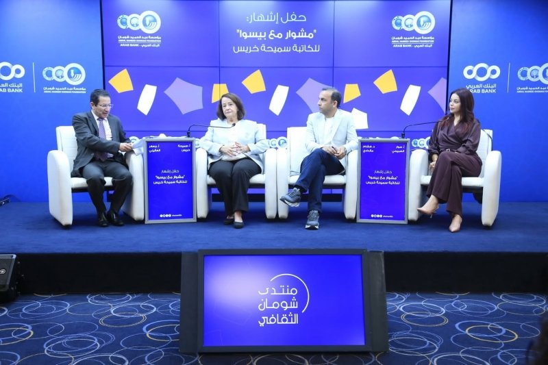 6a5f3d18aec14 Four panelists sit in white armchairs on a blue stage for a discussion, with Arabic banners and logos on the backdrop and small lectern signs in front of each speaker.