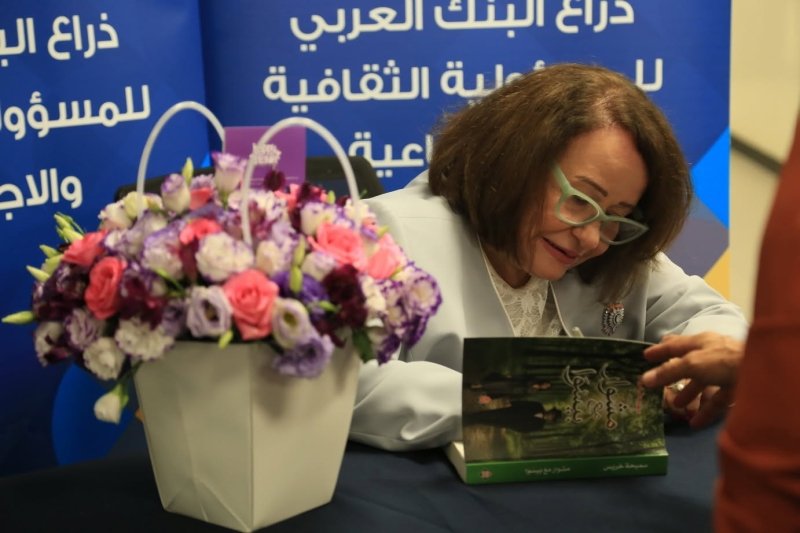 6a5f3d18cc8f6 Woman wearing light green glasses signs a book at a table, beside a white vase of pink and purple flowers during a book signing event.