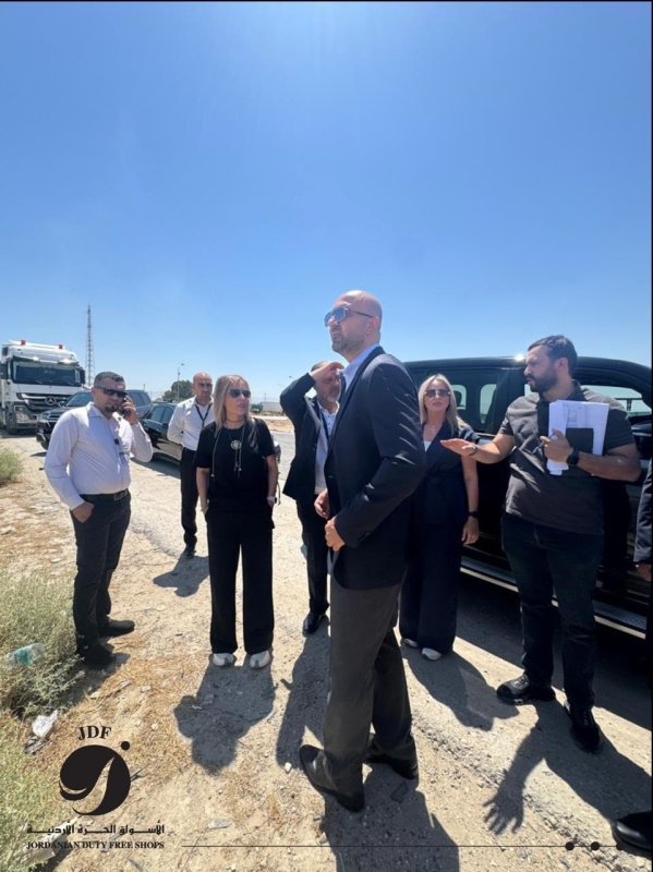 6a607062f35e8 - عروبة الاخباري | Oroba News Group of professionals on a sunny roadside briefing; a man in a suit speaks while others listen beside parked cars and a truck in the background.