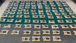 6a631e2acbd1c - عروبة الاخباري | Oroba News Many small beige fabric squares with a dark circular emblem laid out in neat rows over a teal surface in a large workshop.