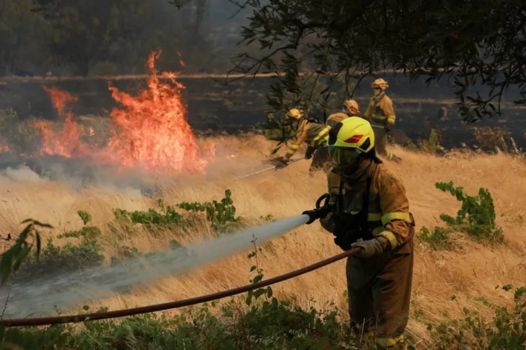 Firefighters in yellow protective gear spraying water on a grass fire in a dry field, with flames and smoke in the background