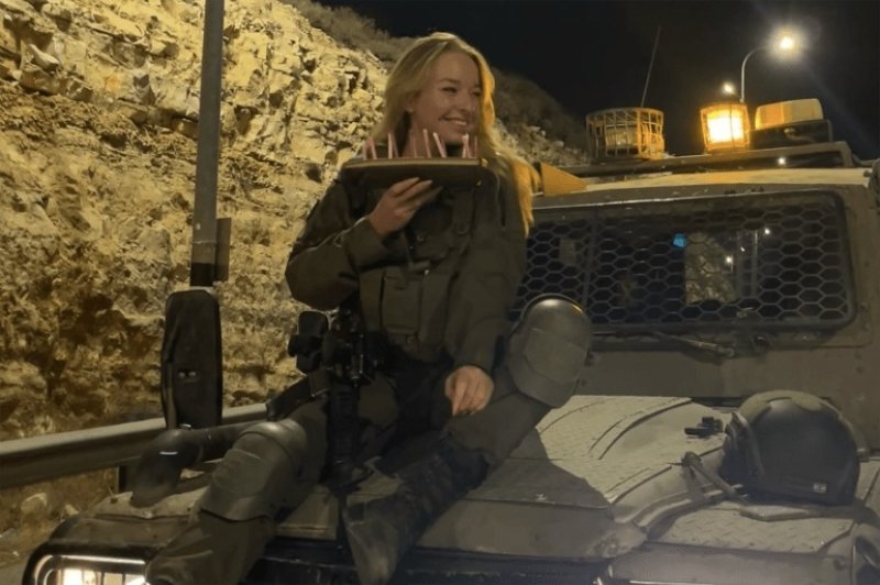 6a6a8c3c45197 Woman in tactical gear sitting on the hood of an armored vehicle at night, smiling and holding a clipboard/photo board.