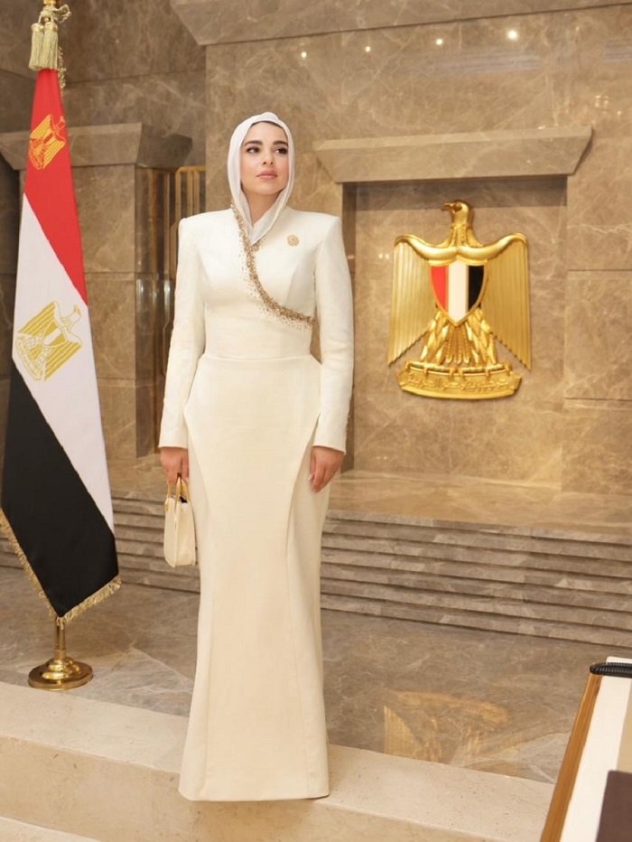 731015650_18408835561159687_6088369334224377741_n_161952 - عروبة الاخباري | Oroba News A woman in a white floor-length gown and hijab stands in a marble ceremonial space beside a gold emblem and the Egyptian flag on a flagpole behind her, conveying formal official attire and setting in Egypt.