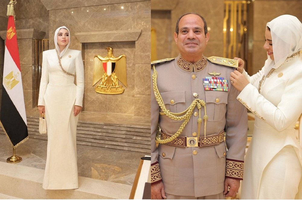 731736916_18408835570159687_888013443035511692_n_162050 - عروبة الاخباري | Oroba News Woman in a white gown stands beside an Egyptian flag and a gold emblem on a marble wall at a formal ceremony.
