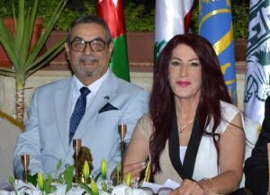 Two adults sit at a ceremonial table with white tulips and gold trophies during a formal event, with flags in the background.
