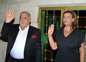 Two formally dressed adults wave hello at the camera: man in a dark suit with white shirt and red pocket square, woman in a black dress, standing by a gold-grilled window.