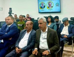 Formal meeting in a conference room with suited men seated; a screen displays two portraits and Arabic text in the background.