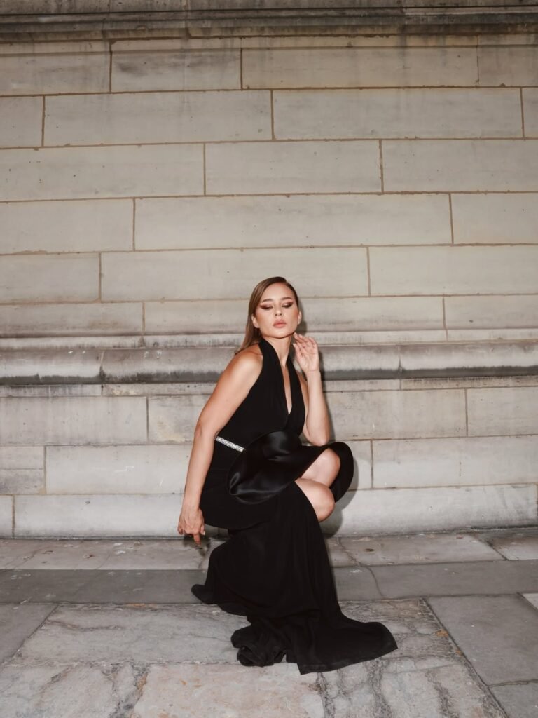 Woman in a black evening gown posing on stone steps, leaning on one knee against a pale stone wall.