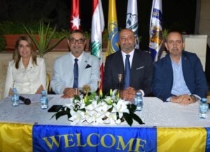 Four suited individuals sit side by side at a decorated table during a formal welcome event, with flags and a flower arrangement in the background.