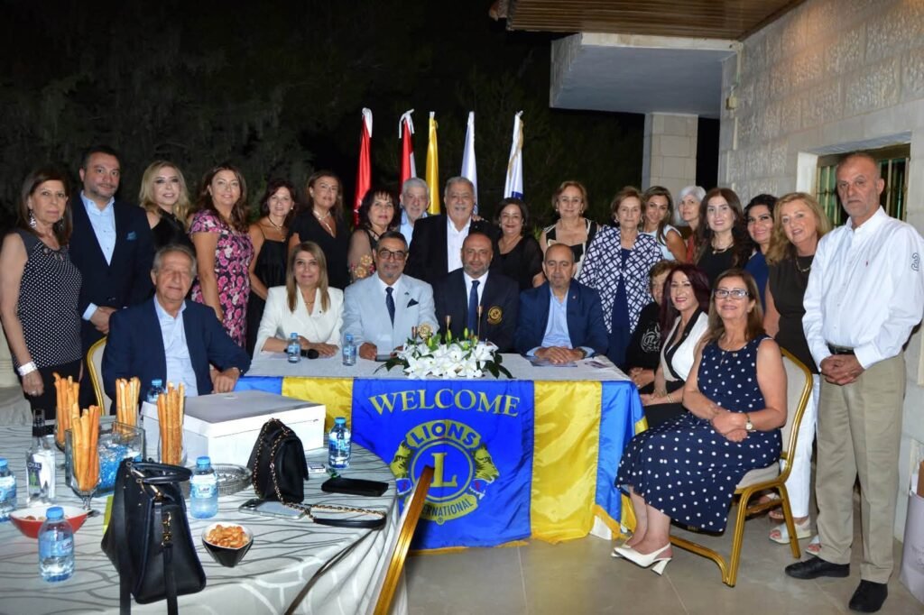 Group of people posing for a photo at a Lions International welcome event, seated and standing behind a table with a blue‑yellow banner and flags in the background.