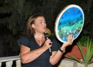 Woman speaking into a microphone while presenting a large decorative clock plate outdoors at night.