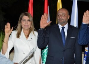 Two professionally dressed adults raise their hands in a ceremonial gesture, standing before national flags.”,