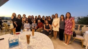 Group of women posing together on a terrace at sunset, with a round table in the foreground and city buildings in the background.