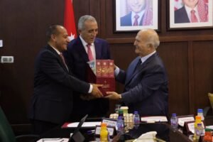 Three men in suits exchange a red boxed set of Arabic books during a formal meeting, with flags and portraits in the background.